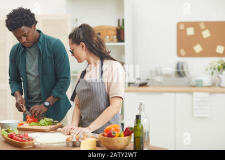 Giovane africano che taglia le verdure per l'insalata lui che aiuta la moglie che fa l'impasto che preparano la cena insieme nella cucina Foto Stock