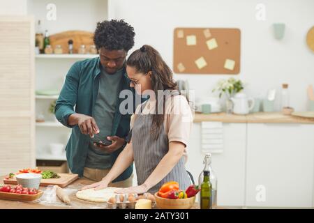 Giovane africano che mostra la ricetta nel suo telefono cellulare a donna che fa pasta per la pizza in cucina a casa Foto Stock