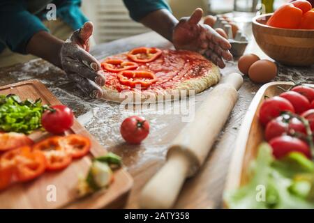 Primo piano di uomo africano che fa la pizza con verdure al tavolo di legno in cucina Foto Stock