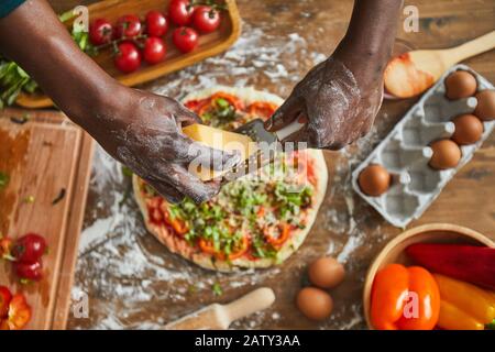 Primo piano di uomo aggiungendo formaggio sulla sua pizza fatta in casa mentre cucinava al tavolo di legno in cucina Foto Stock