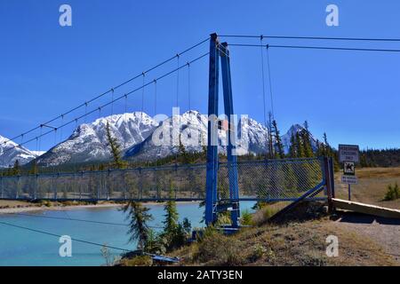 Ponte sospeso blu sul fiume turchese. Alte montagne coperte di neve, cielo blu, giornata di sole. Kootenay Pianure Riserva Ecologica, Canada. Foto Stock