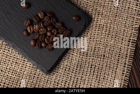 Mucchio di chicchi di caffè tostati su vassoio in pietra nera su sacco di hessian e superficie in legno Foto Stock