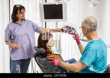 Team medico dentista in studio dentale parlando con paziente femminile e preparazione per il trattamento. Il dentista senior mostra l'immagine dei raggi X al paziente Foto Stock