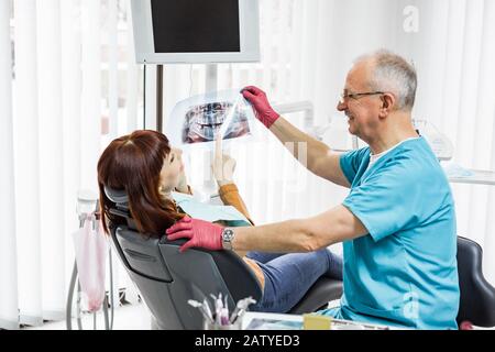 Sorridente dentista professionista di sesso maschile che mostra raggi X dentali a paziente giovane donna, seduto in sedia dentale in moderna clinica dentale Foto Stock