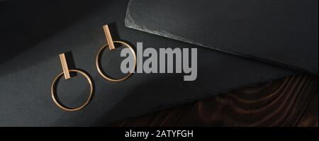 Vista dall'alto degli orecchini dorati dal design su lastre di pietra nera su sfondo di legno Foto Stock