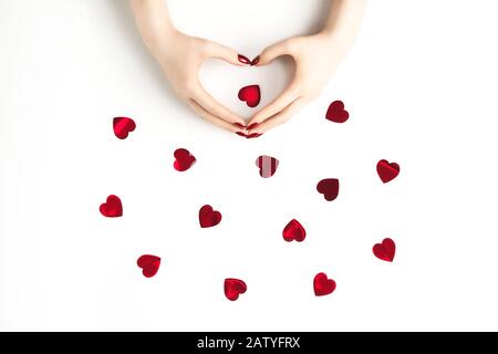 Belle mani femminili con vivaci confetti rossi scintillanti a forma di cuore versati su sfondo bianco isolato. Sfondo per il tuo design. Creativa alla moda Foto Stock