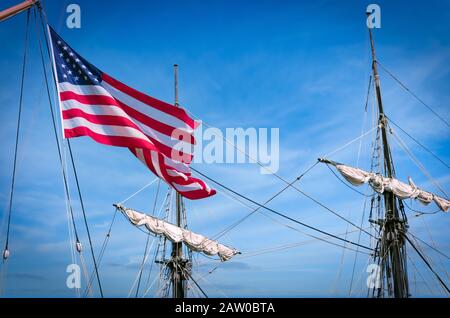 La bandiera americana soffia nel vento con alberi di nave e vele sullo sfondo. Foto Stock