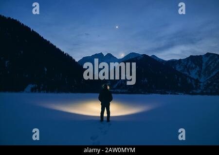 Silhouette dell'uomo illumina il lago ghiacciato con la lampada frontale sulle montagne sotto il cielo notturno con stelle e luna. Foto Stock