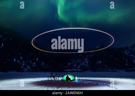 Uomo in campeggio con bicicletta e tenda verde al lago ghiacciato in montagna con aurora boreale. Drone che illumina il cerchio di luce sopra il campo. Foto Stock
