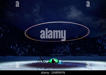 Uomo in campo con bicicletta e tenda verde al lago ghiacciato in montagna. Drone che illumina il cerchio di luce sopra il campo con starry notte cielo backgroun Foto Stock