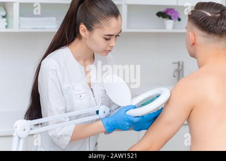 Un dermatologo esamina la pelle sul corpo di un giovane uomo attraverso una lente di ingrandimento di una lampada di cosmetologia. Trattamenti di bellezza Foto Stock