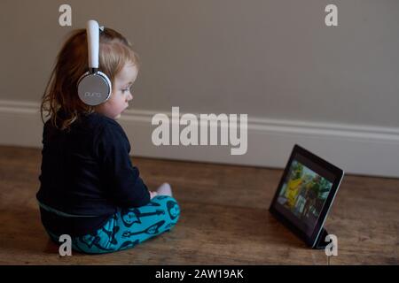 Ragazza di 15 mesi che indossa le cuffie e guarda un film su un iPad, Regno Unito Foto Stock