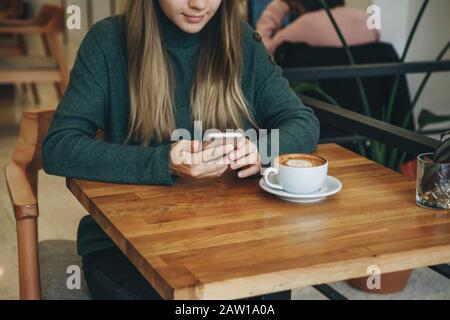 La ragazza utilizza un telefono cellulare e beve caffè. Ella ha una riunione o beve la mattina con un cappuccino o un piatto bianco. Foto Stock