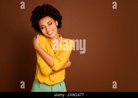 Foto di affascinante bella pelle scura signora tenendo le spalle occhi chiusi godere di amore se stesso bella blouse vestiti indossare camicia gialla pantaloni verdi Foto Stock