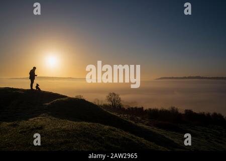 Un uomo e il suo cane ammirano l'inversione della nuvola sulla Città di Bath nel Somerset all'alba da Solsbury Hill in un giorno d'inverno. Foto Stock