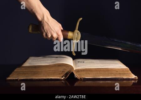 Mano maschio forte con la spada sopra la Bibbia aperta Foto Stock