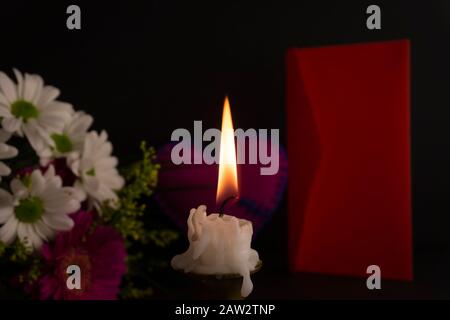 Candela corta che brucia sul pavimento in primo piano tra i fiori, accanto alla busta rossa contro sfondo scuro. Concetto romantico di tema serale Foto Stock