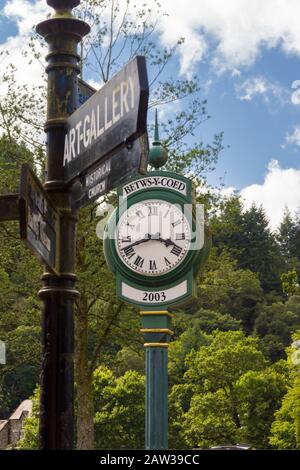 L'orologio del villaggio con un cartello fuori dalla stazione ferroviaria che punta a vari punti di interesse nella città di Betws-y-Coed North Wales Foto Stock