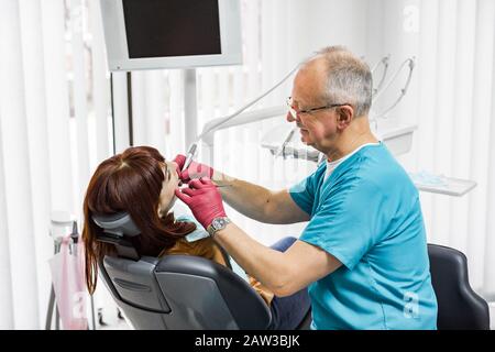 Vista laterale della paziente femminile esaminata dal dentista maschile senior, indossando guanti in lattice, in una moderna clinica dentale. Foto Stock