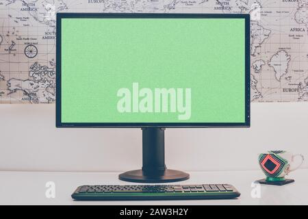 Computer desktop vuoto con tastiera e tazza di caffè su tavolo bianco in sala soleggiata, mock up. Sfondo della mappa del mondo. Schermo verde. Stile retrò. Foto Stock