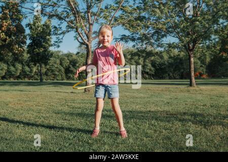 Cute sorridente caucasica prescolare ragazza giocare con hoola hoop nel parco all'esterno. Attività sportiva per bambini. Stile di vita felice infanzia. Estate stagionale all'aperto Foto Stock