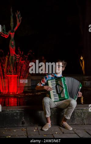 Kaunas, Lituania - 19 05 2013: Giovane musicista di talento inidentificato che suona la fisarmonica nel vicolo della libertà in fontana illuminata e sculpt della dea del mare Foto Stock