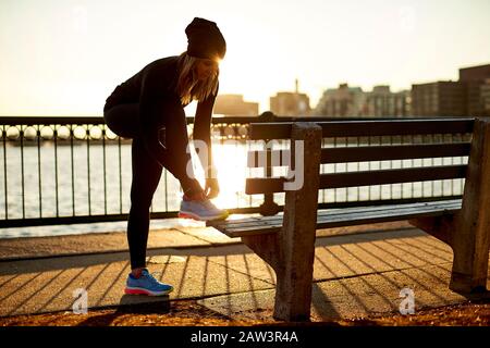 Una donna atletica con retroilluminazione lega la sua sneaker a una panca da parco. Foto Stock