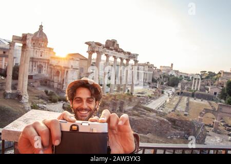 Giovane uomo turistico zaino in spalla che prende selfie con fotocamera d'epoca al Foro Romano all'alba. Storico Foro Romano imperiale a Roma, Italia da Foto Stock