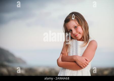 Piccola ragazza bionda leggermente sorridente in abito bianco, ritratto esterno con luce naturale del cielo Foto Stock