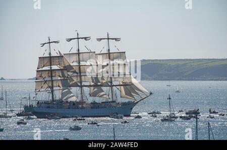 Enorme goletta a tre alberi, retroilluminata al sole, naviga nel vento, scortata da molte piccole barche e yacht fuori Falmouth come parte della Tall Ships Race Foto Stock