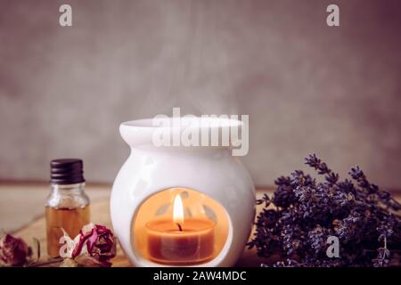 Immagine in stile vintage di candela bianca in ceramica aroma lampada ad olio con bottiglia d'olio essenziale e petali di fiori secchi su disco di legno di pino naturale, fondo secco Foto Stock
