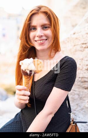 Felice positivo giovane zenzero femmina guardando la macchina fotografica mentre si siede con cono gelato sulla strada della città e godendo la giornata estiva Foto Stock