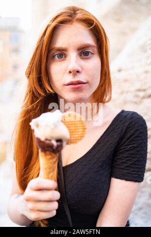 Calmi la giovane femmina rossa in abito casual che tiene cono gelato con salsa di cioccolato e biscotto e guardando la fotocamera Foto Stock