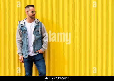 Ritratto di un giovane uomo freddo su sfondo giallo. L'uomo indossa jeans blu, una giacca in denim e occhiali da sole. Foto Stock