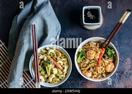 Due ciotole di tagliatelle thailandesi con curry, gamberetti e verdure su una superficie di legno con salsa di soia e una stuoia di bambù Foto Stock