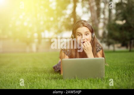Buone notizie. La donna entusiasta stupita sorpreso da ciò che vede sul suo portatile sdraiato sul prato verde erba fuori nel parco della città cuffie in testa Foto Stock