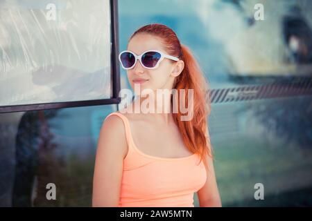 La giovane donna redhead seduto alla fermata dell'autobus si aspetta l'autobus all'aperto. Elegante ragazza in camicia rosa casual, in vetro da occhiali a forma di farfalla Foto Stock