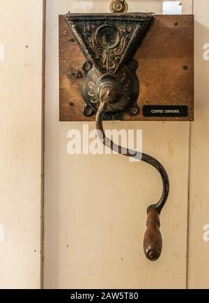 Waimea, Hawaii, Stati Uniti. - 15 Gennaio 2020: Sede Centrale Di Parker Ranch. Macinacaffè in legno di metallo marrone vecchio con manico lungo fissato a parete verniciata beige. Foto Stock