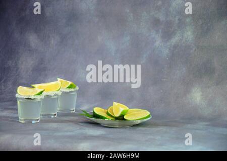 Tre bicchieri di tequila di vetro con sale ai bordi e fette di calce si ergono su un tavolo di legno accanto ad un piatto di calce tagliato da anelli con un pizzico di