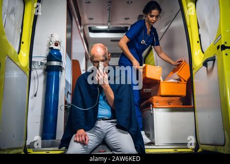L'uomo respira attraverso la maschera di ossigeno, l'infermiera giovane apre il sacchetto medico nell'automobile dell'ambulanza Foto Stock