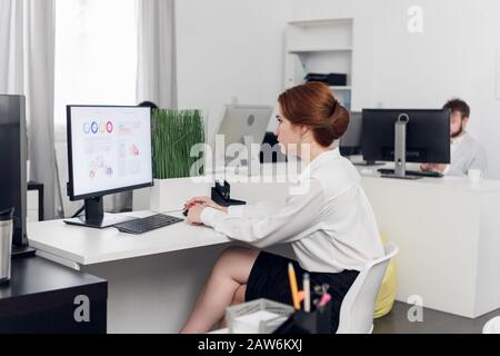 Giovane donna nella gonna si siede a un tavolo e guarda sullo schermo del computer in ufficio Foto Stock