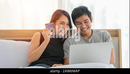 Asian Coppia shopping online e pagamento con carta di credito al computer portatile, coppia Felice a casa navigare la rete a letto Foto Stock