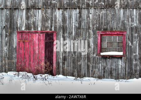 Primo piano di vecchi fienile di tavole di legno grigio in un campo di neve con porta rossa e finestra rossa Foto Stock