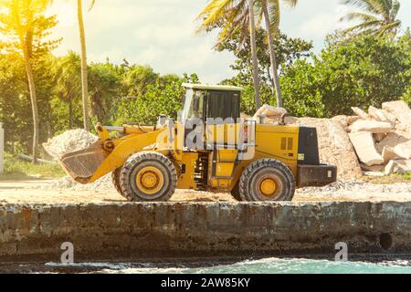 Grande bulldozer giallo con martello di sollevamento sul sito di costruzione sullo sfondo di palme. Costruzione di un hotel sull'isola. Foto Stock