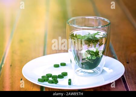 Clorofilla in un bicchiere d'acqua e pillole verdi di spirulina su un piatto su tavola di legno. Spazio di copia, luce solare. Foto Stock