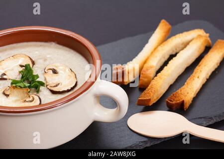 Zuppa di crema di funghi fatta in casa con funghi a fette in ciotola di porcellana, cucchiaio di legno e toast su una tavola di pietra nera. Foto Stock