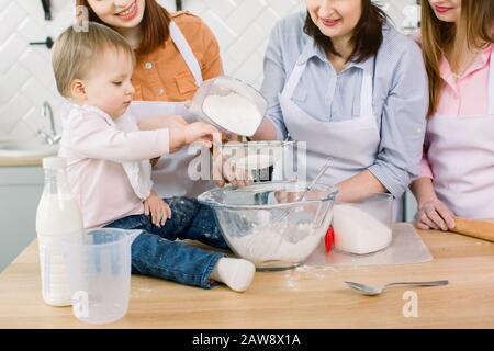 Adorabile bambino con madre, zia e nonna che fanno l'impasto con farina e uova e zucchero insieme a casa. Donne in grembiuli bianchi e cappelli da chef pista Foto Stock