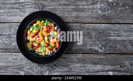 insalata di spinaci con gamberi avocado e pomodori secchi, formaggio sbriciolato, cipolla rossa in una ciotola nera su un rustico tavolo di legno vista orizzontale dall'alto, Foto Stock