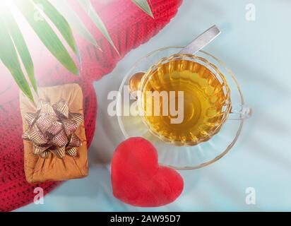 Tè mattutino di San Valentino. Bicchiere di tè trasparente in vetro, morbido cuore rosso, scatola regalo avvolta nella tecnica Furoshiki, ponticello a maglia rossa su dorso blu Foto Stock