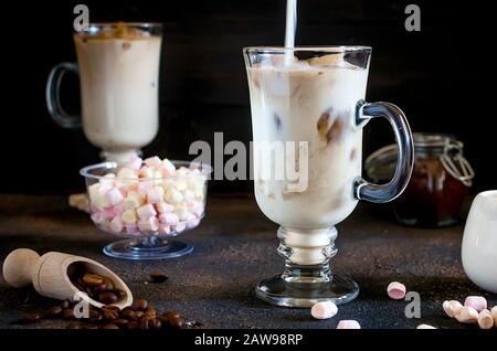 Caffè estivo Iced in bicchieri alti fatti con cubetti di ghiaccio da caffè, latte o crema versato sopra e marshmallows decorato, sfondo marrone, selettiva foc Foto Stock